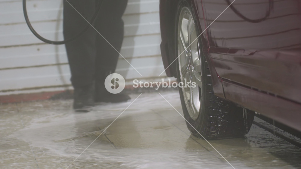 low angle close up of person wearing black pants and canvas shoes rinsing red car exterior with high pressure water spray inside indoor