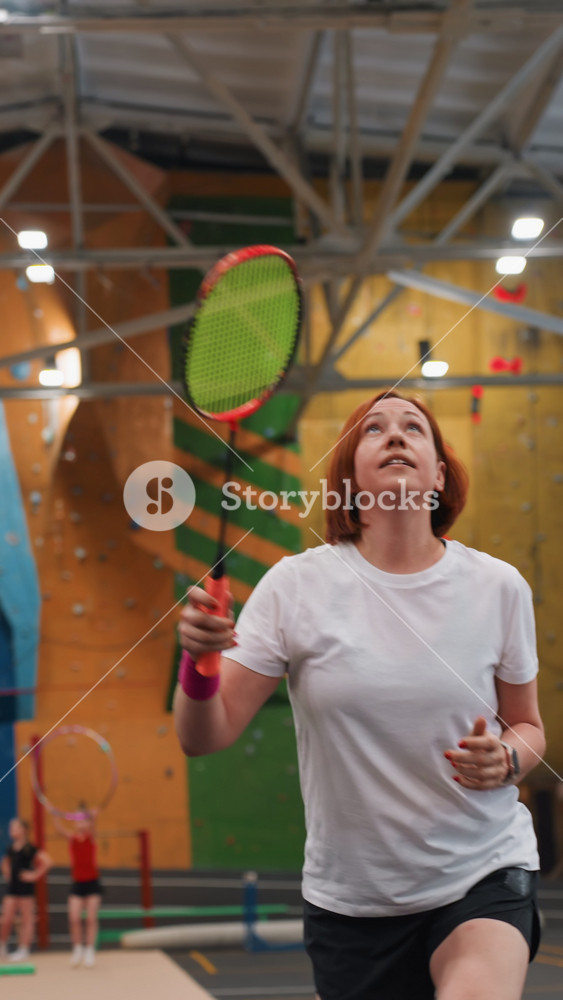 Relaxed woman warming up at gym court. Friendly woman enjoying light badminton drills inside gym. Casual woman engaging in indoor badminton