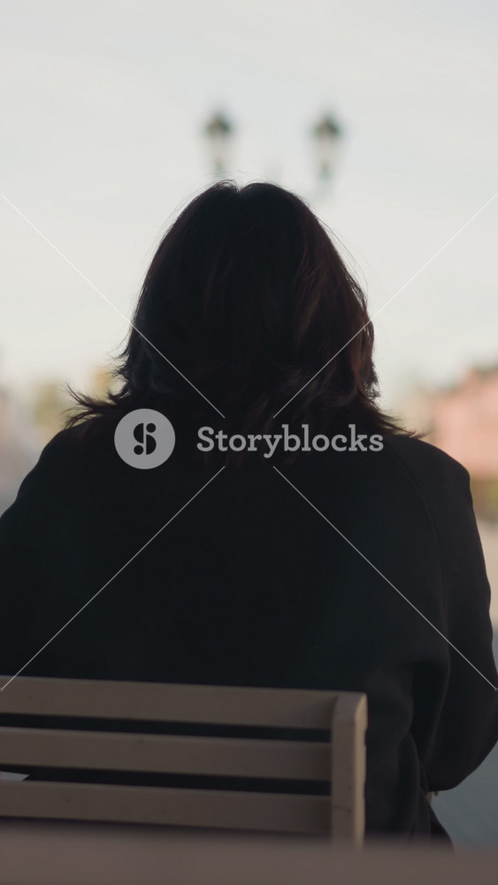 Rear view of woman seated outdoors on wooden chair in urban setting, wearing black coat with soft natural light, blurred background of