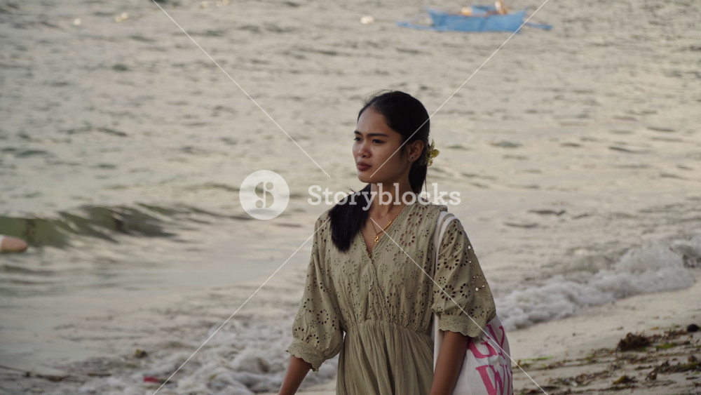 Young asian woman gazing beach horizon at dusk, barefoot at waterline, colorful outrigger boat nearby, floral hair clip, tote on shoulder