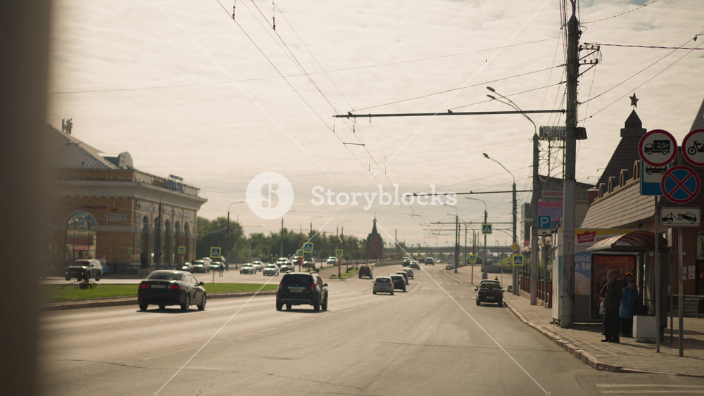 Wide city street with cars, traffic signs, and streetcar wires, showing urban environment. People wait at bus stop near buildings on sunny