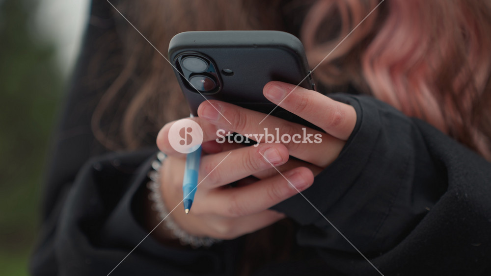 student holding phone and pen, closeup of female hands with pink hair and pearl bracelet wearing black hoodie, outdoor blurred green