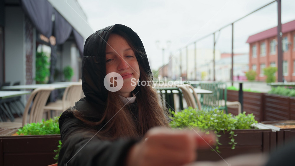 hooded woman greeting friend outdoors, reaching hand and playful block, paper cup held, terrace plants and urban street backdrop, candid