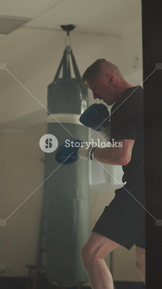 Athlete hits punching bag by fists and legs in gym. Muscular man exercises kick on equipment in fighting club slow motion. Martial arts