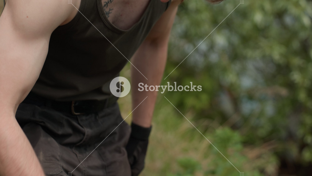 Close-up waist view of soldier brushing dirt off gloves and cargo pants after crawling, with muscular arms exposed in sleeveless top