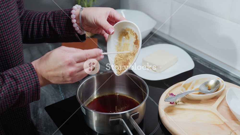 Partial view of person in checkered sleeve pouring powdered ingredients into hot oil pot on electric stove, with wooden tray, spices, and