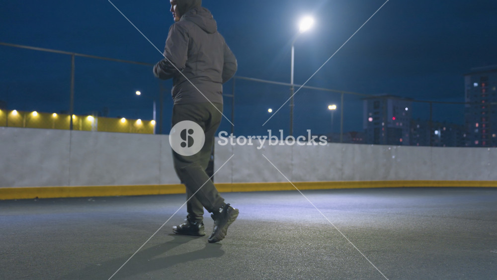 Partial view of athlete kicking soccer ball toward goal post during night training session on urban outdoor field, city lights and buildings