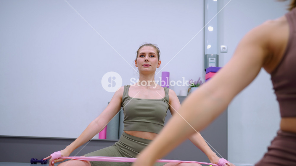 Close-up of a woman in brown engaged in a fitness exercise, stretching with a ribbon in a gym, alongside another woman in green, with