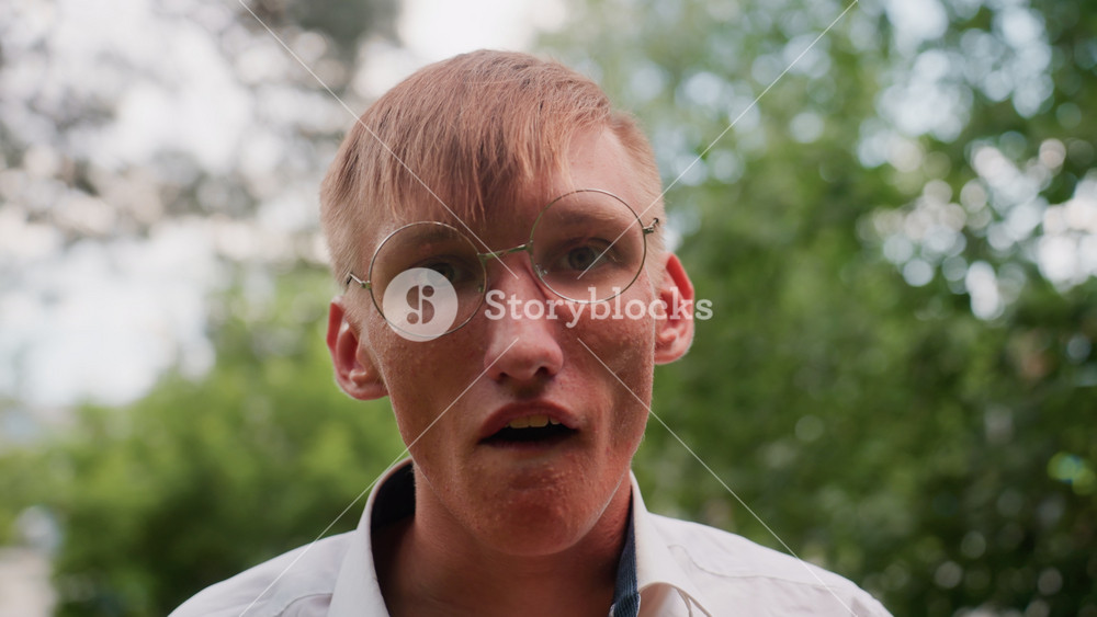 Close up view of young man in white shirt with glasses walking tired in blurred green forest background, touching face and showing signs of