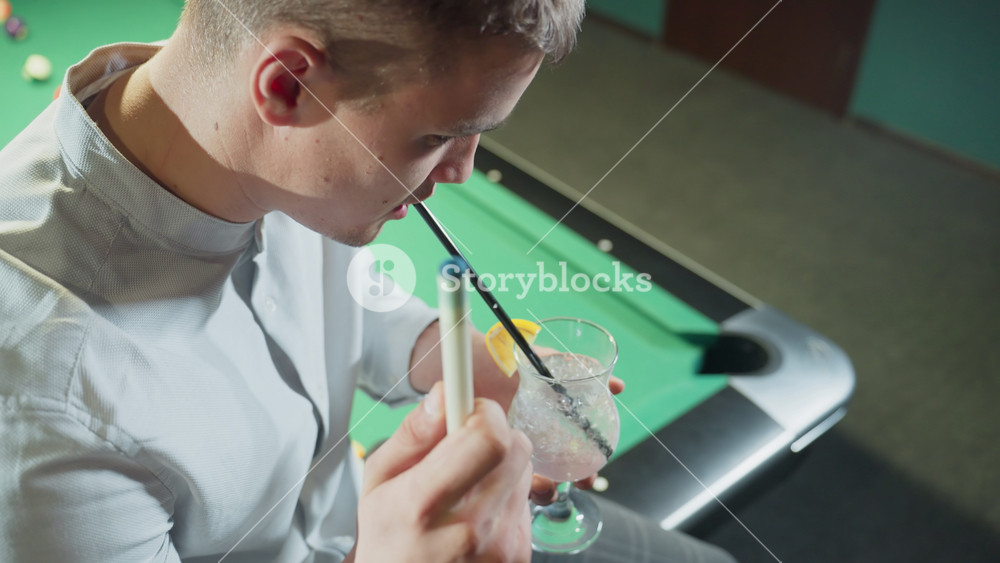 Close-up of young man wearing white shirt and grey trousers, sitting on green pool table holding cue stick and of lemon drink with black
