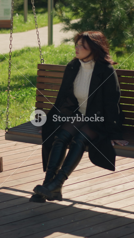 Young woman in stylish black coat and boots sits on a wooden swing in a sunlit park, gazing serenely into distance, surrounded by lush