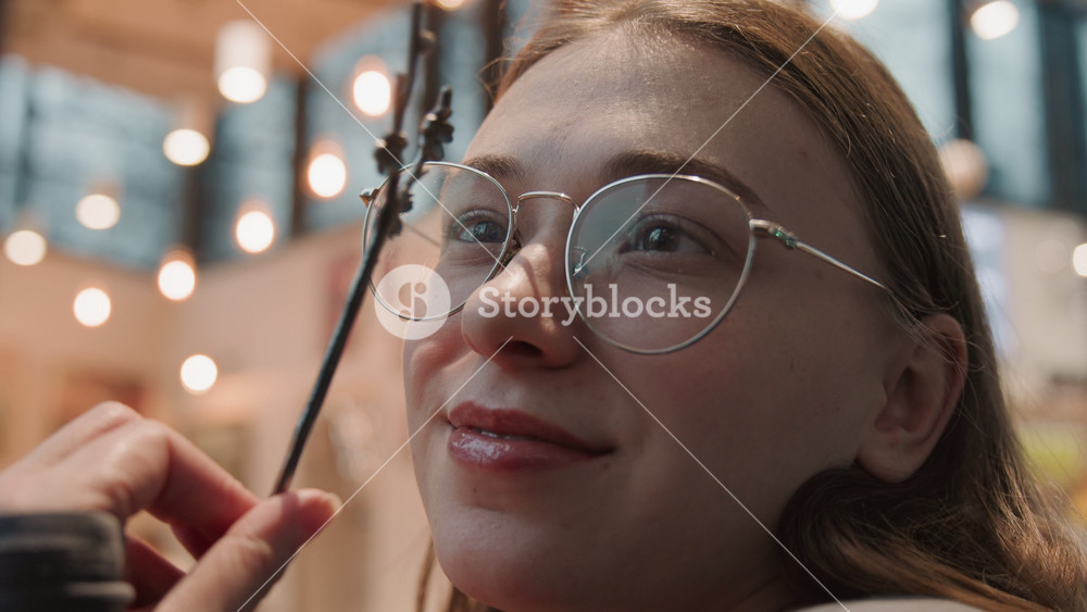 Young white woman holding blossom branch in cafe, playful touch near glasses and cheek, warm bokeh lights and whimsical mood, soft smile and