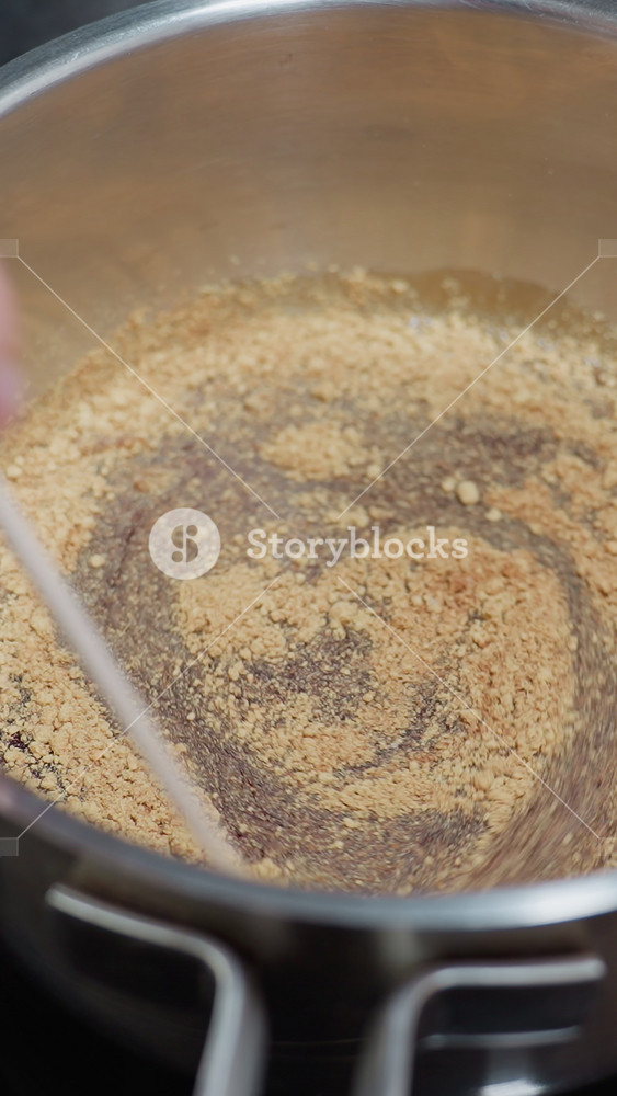 Close-up of hand using a stick to stir soup in stainless steel pot on electric stove, with powdered ingredients mixing in liquid, wooden