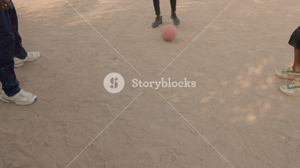 Black boys playing street soccer dusty ground, closeup of pink ball and feet as young defenders and dribbler clash sneakers kick up sand and