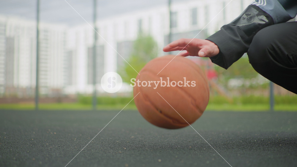 closeup urban basketball dribble on asphalt, hand guiding ball while kneeling in jacket near fenced city court with apartment buildings in