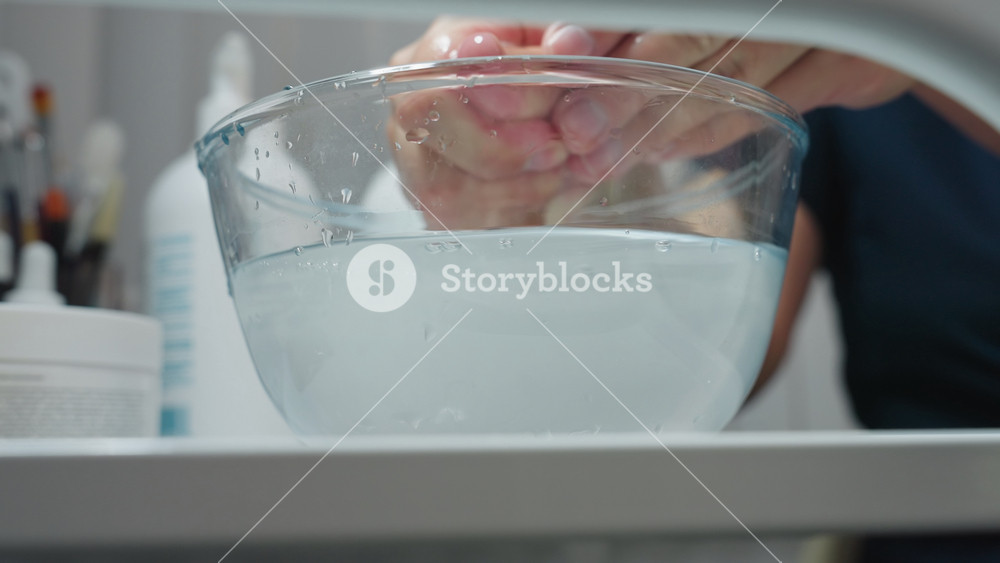 Hand view of facialist dipping cotton pad into glass bowl of purified water on white trolley beside skincare bottles, then gently pressing