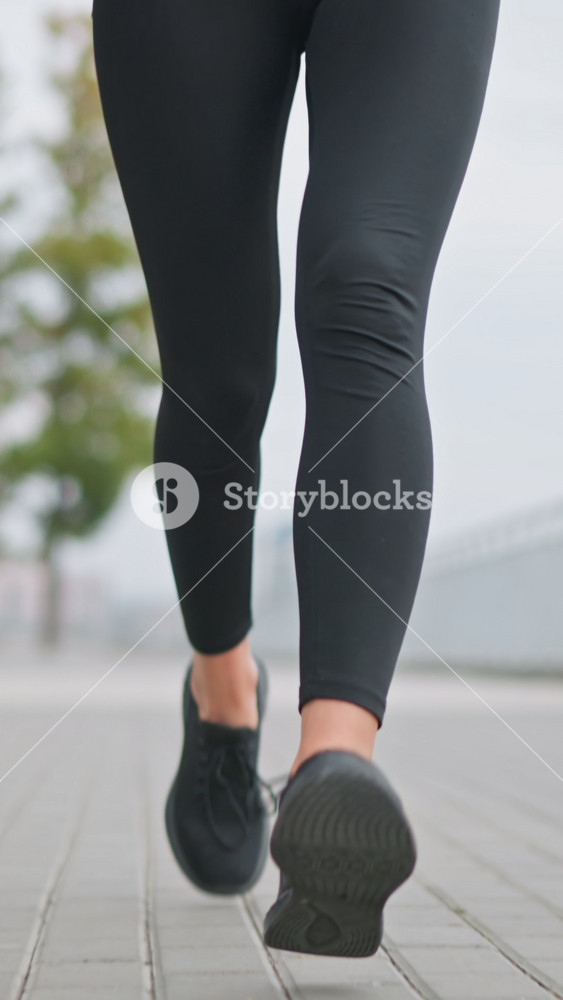 Close-up of woman s legs in black leggings and sneakers jogging in outdoor path with blurred background of trees, poles, and iron fence