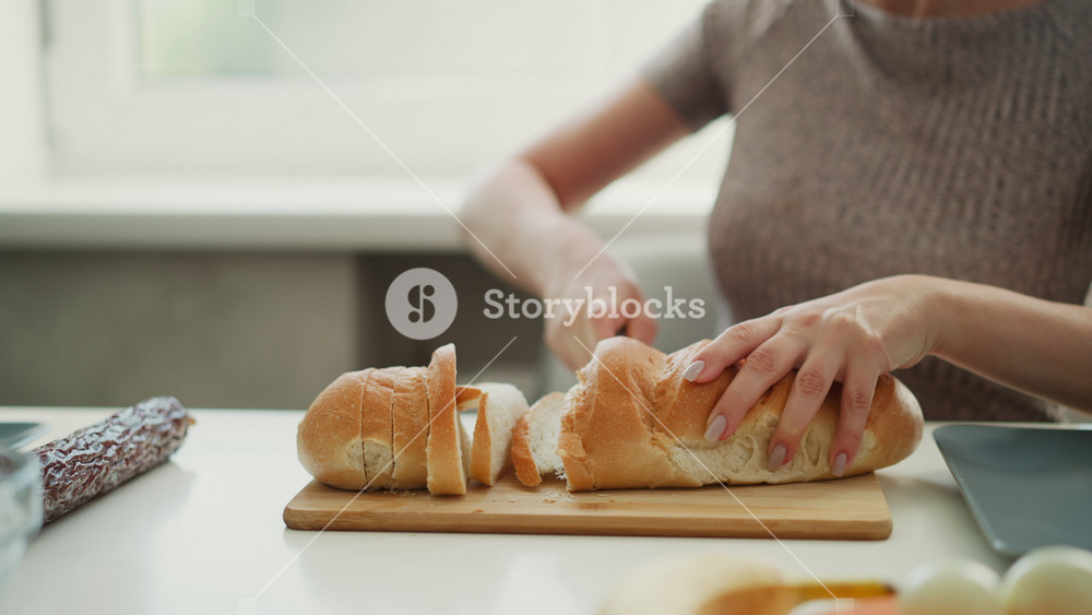 Close up of young woman with manicured nails firmly holding loaf of bread slicing carefully on cutting board next to eggs and plates on