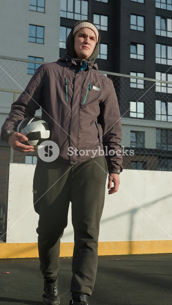 Footballer in hoodie and beanie holding ball in right hand walks along football pitch with goal post, tall residential building in