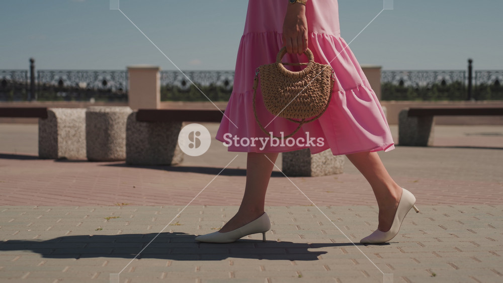 pink dress woman walking on promenade, closeup of legs and straw bag, white heels, stone benches and flower planters, warm sunlight casting