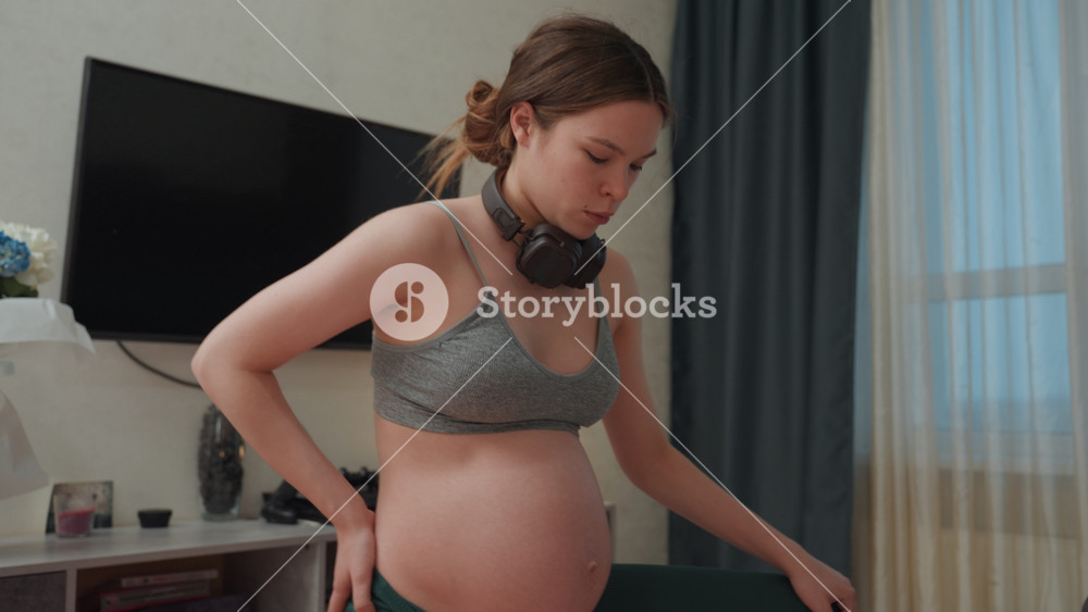 Pregnant woman stretching at home, lightfilled bedroom scene showing sports bra and leggings, hands on lower back for support, headphones