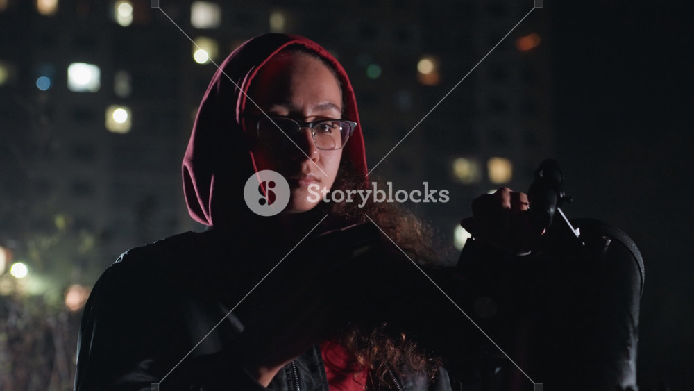 rooftop night stargazer checks telescope and binoculars under city lights, hooded woman with glasses adjusts scarf and equipment moody