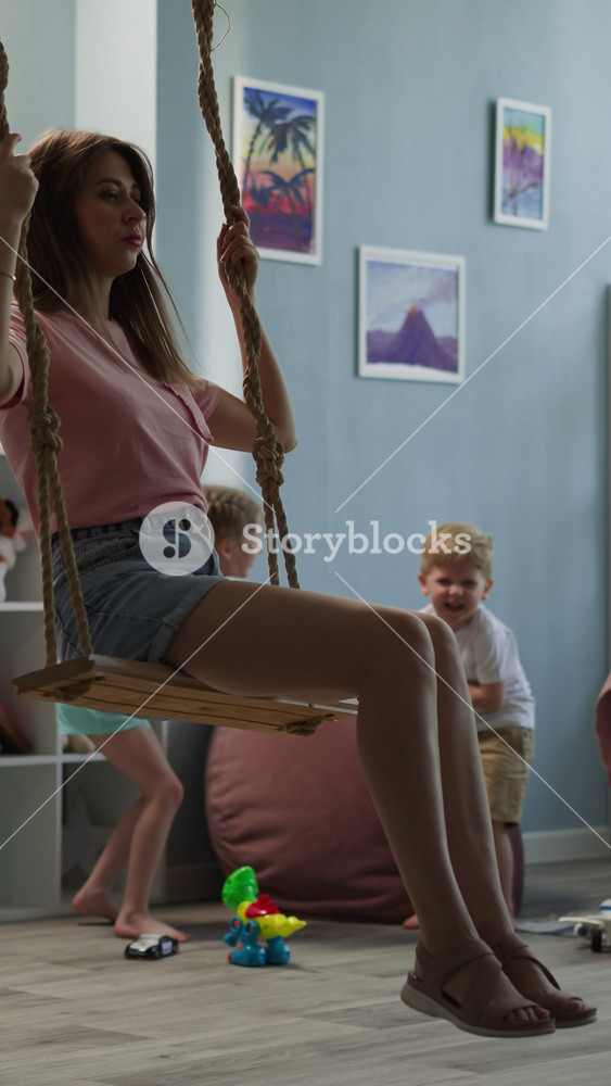 Young mother rests with kids at home. Woman rides swing calmly while daughter and son play with toy swords. Happy family has fun together