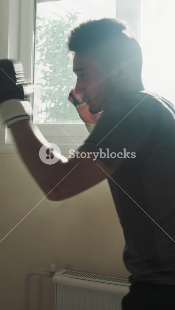 Athlete attacks punching bag with fists and leg in sports club. Young kick-boxer kicks and punches heavy equipment in gym at back sunlight