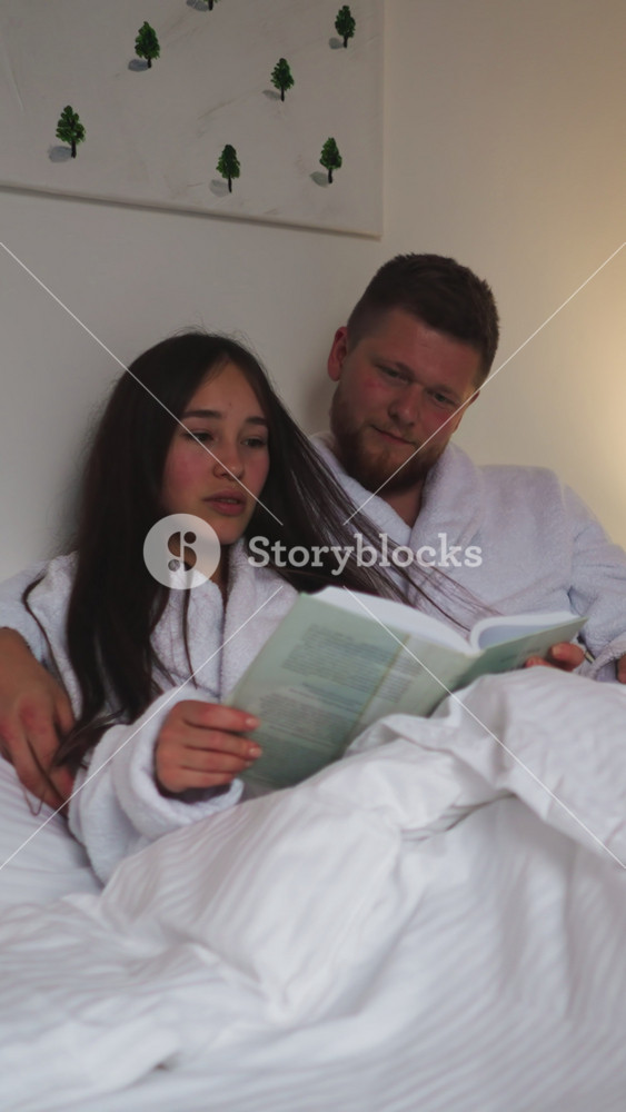 Man caresses loose hair of wife reading book in bed. Relaxed couple in soft bathrobes enjoys reading novel in bedroom at night. Romantic