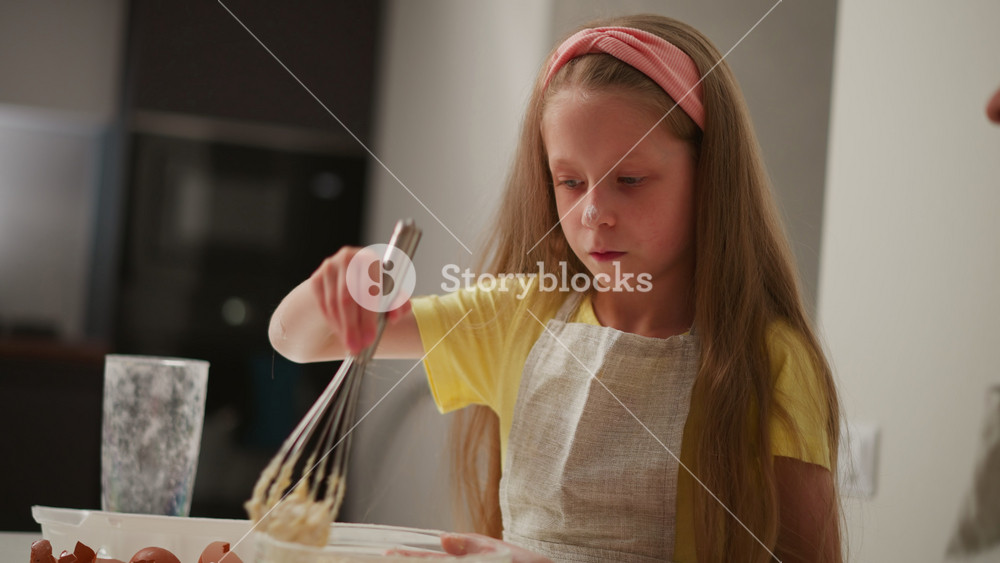 close up of young girl in yellow shirt and apron whisking flour in bowl as adult supports bowl from below, broken eggshells scattered on