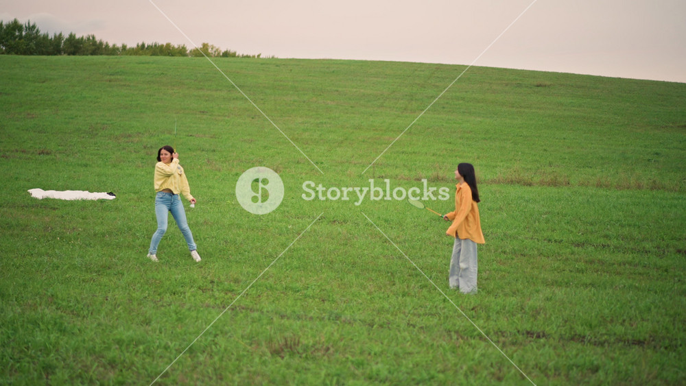 Asian women meet in green field across wide grassy hill, approaching near small white blanket, casual jeans and yellow tops, cinematic
