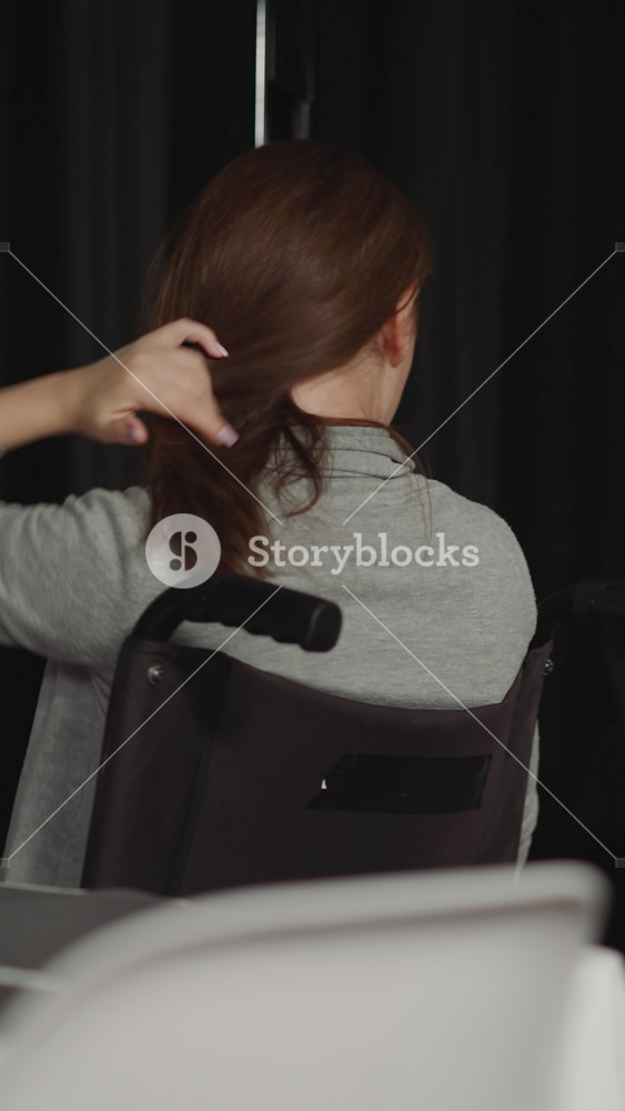 Young woman moves to window in wheelchair adjusting hair with hand. Lonely housewife with spinal cord injury sits in kitchen staying late