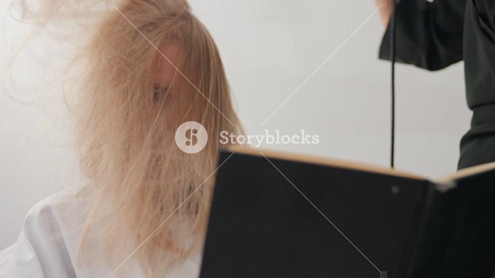 Frizzy blonde hair covering face, caucasian woman sits in salon cape while stylist lifts dryer closeup back view shows tangled strands and