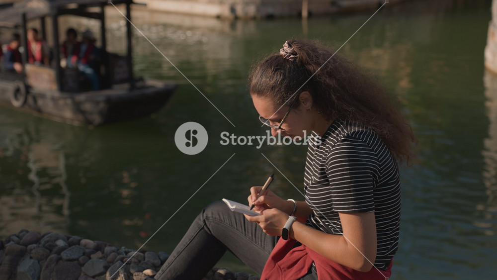 Woman sketching near tranquil water. Young woman designing intricate art beside serene canal setting. An aspiring artist creates detailed