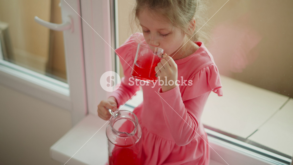 close up aerial view of kid wearing pink dress seated by window pouring red juice from jar into clear mug spilling drink on cloth indoors