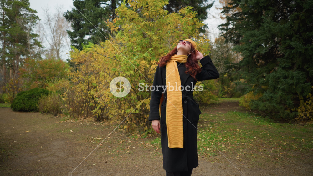 Elegant woman in black coat with yellow beret and scarf stands in autumn park, looking around and touching her red hair with a warm smile