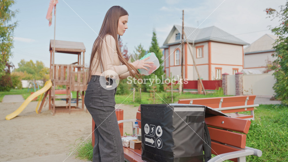 white woman unpacking delivery bag outdoors, arranging groceries from insulated delivery bag on park bench near playground and suburban