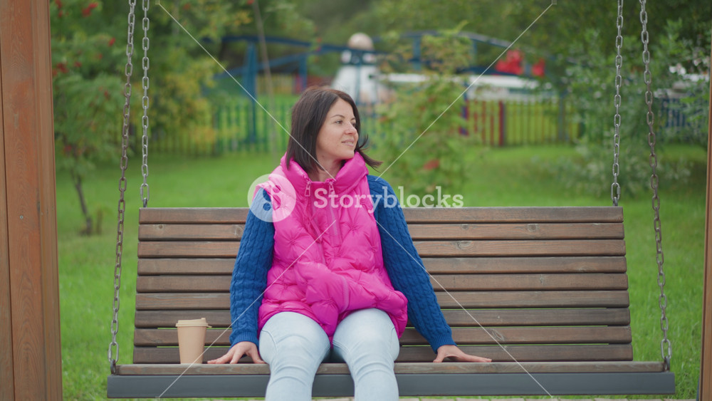 woman on wooden swing bench, wearing bright pink vest and blue sweater, coffee cup on seat, bicycle parked nearby, green park background