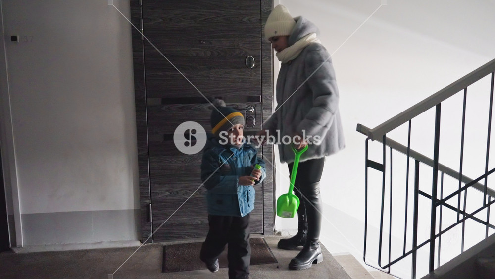 Winter stairwell mom and son enter apartment door, mom hands keys while child holds toy shovel, bundled coats, hat and scarf, quiet stair