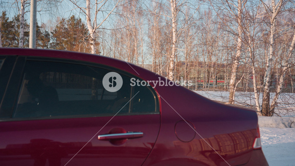 Man in hoodie shuts trunk of red car and walks backward in snowy lot with bare trees in background and faint outline of another car visible