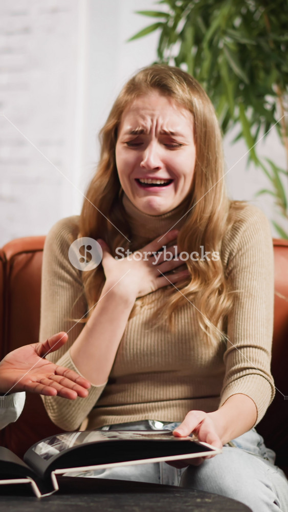 Young woman laughs surprised of African-American husband childhood pictures in photo-album sitting on couch in living room slow motion