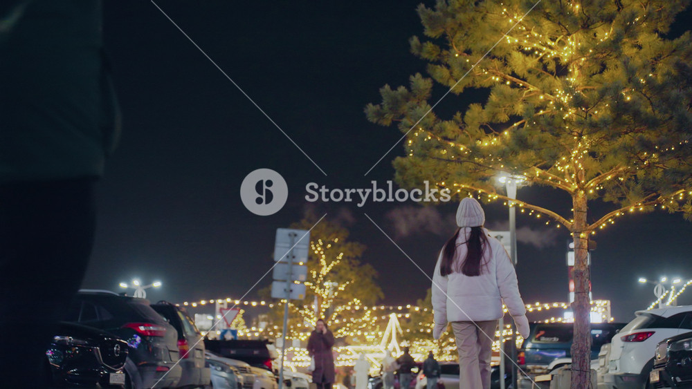 Back view of woman wearing winter outfit and beanie walking through bustling city street at night, surrounded by glowing festive lights
