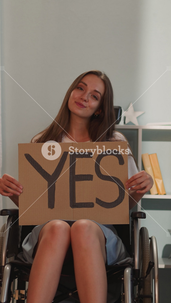 Positive brunette woman turns poster with word Yes to camera leaning head to side. Young person with disability sits in wheelchair smiling
