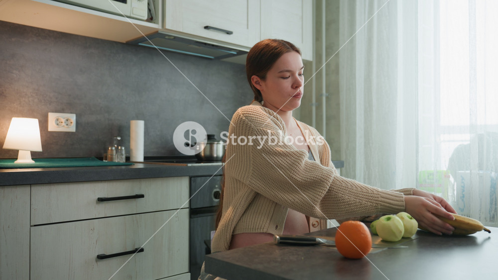Pregnant white woman in kitchen holding orange and apples, wearing cozy cardigan and bralette, exposed baby bump, soft lamp glow and morning