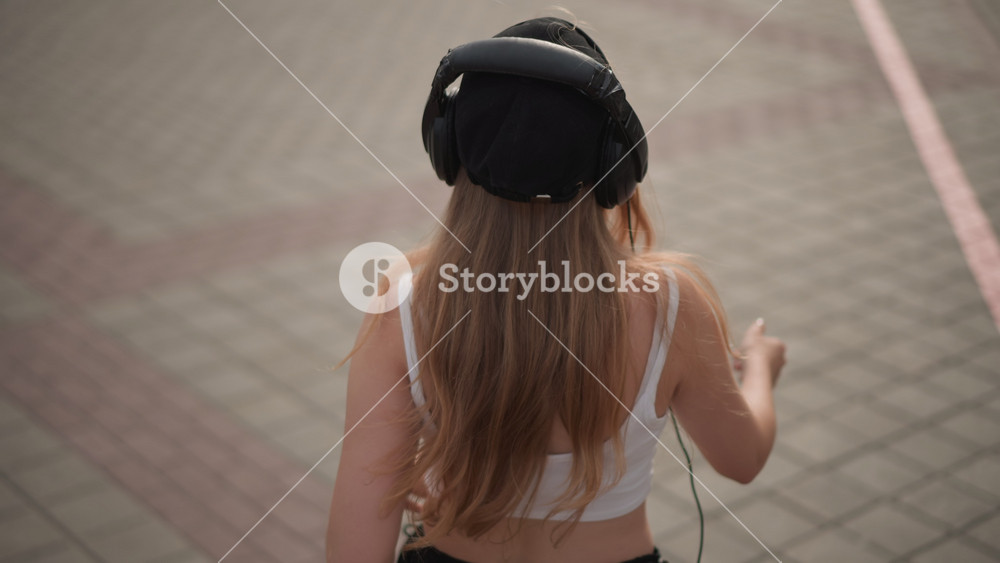 young woman on street wearing headphones adjusting beanie and hair, dancing casually on tiled plaza, warm daylight, candid closeups