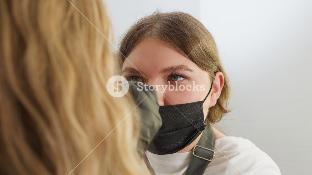Beautician wearing black face mask carefully applies small amount of cream to blonde client's face during eyebrow grooming session in bright