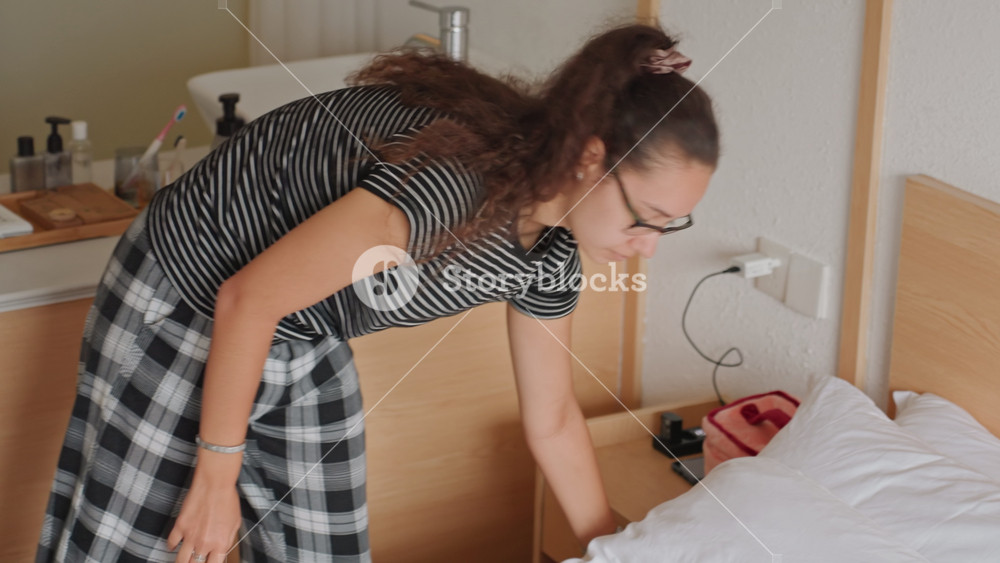Hotel room guest making bed, casual morning scene with woman in plaid pajama pants smoothing duvet and arranging pillows near wooden