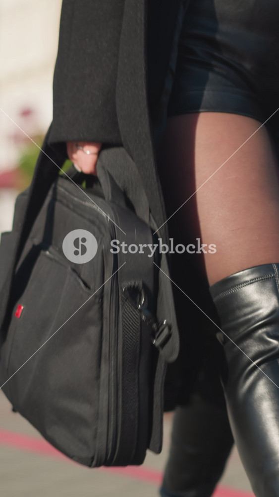 Close-up of stylish woman walking confidently on sunlit urban street, wearing black coat and leather boots, carrying two bags, with leg