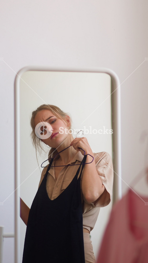 Young confused woman tries on elegant pink and classic black dresses against mirror choosing fashionable outfit for evening event slow