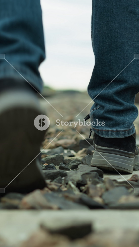 Close-up view of a man's legs, in jeans and canvas shoes, walking in an irregular way on rocky railway tracks, with a blur view of green
